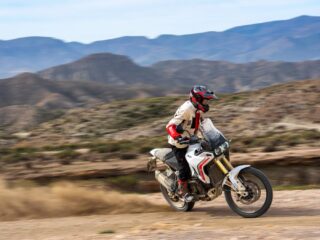 ライダーが白いドゥカティのオフロードバイクで砂漠のような未舗装路を走行している様子