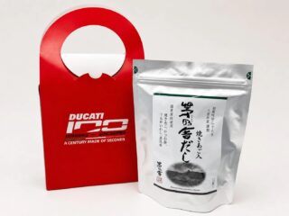 DUCATIの100周年記念ロゴがデザインされた赤い紙袋と、久原本家茅乃舎の「焼きあご入 茅乃舎だし」の銀色のパッケージが並んでいます