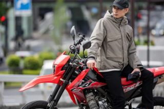 ベージュのジャケットと黒いキャップを着用した男性が、赤いオフロードバイクに腰掛けている