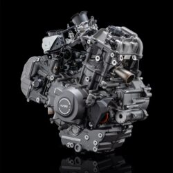 黒い背景に、多くのボルトや配管が見える精密なV型2気筒エンジンがクローズアップで写っています
