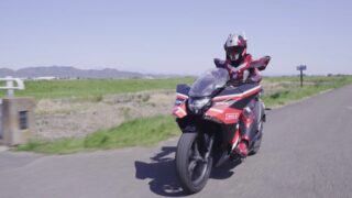赤と黒の未来的なスーツを着た人物が、同じ色のスポーツバイクに乗って田舎道を走行している様子
