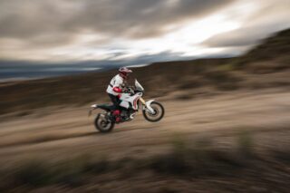 広大な荒野の砂利道を、白いアドベンチャーバイクに乗ったライダーが高速で走行しています